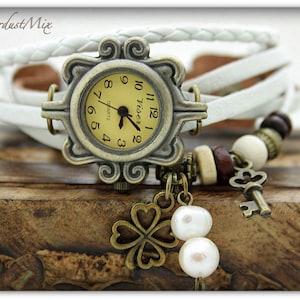 Può includere: Un orologio da polso in pelle bianca con un quadrante dorato e un ciondolo a forma di chiave. L'orologio ha un movimento al quarzo e un ciondolo a forma di quadrifoglio.