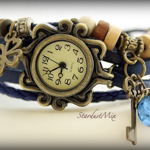 Puede incluir: Un brazalete de reloj de cuero azul con una esfera de reloj dorada, un amuleto de trébol de cuatro hojas y un amuleto de llave con un cristal azul. La esfera del reloj tiene números romanos y la palabra "Quartz".