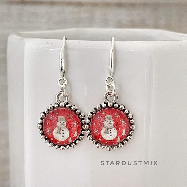 May include: A pair of silver-toned dangle earrings featuring circular charms with a red background. Each charm displays a white snowman illustration with a hat and scarf, surrounded by small white snowflakes. The text STARDUSTMIX is visible.