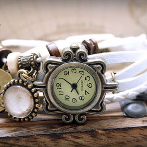 Puede incluir: Un reloj de pulsera de cuero blanco con una esfera redonda de color dorado antiguo. La esfera del reloj tiene marcadores de hora con n&uacute;meros romanos negros y agujas negras. El reloj est&aacute; adornado con un dije que dice "Passionate" y un peque&ntilde;o dije redondo blanco.