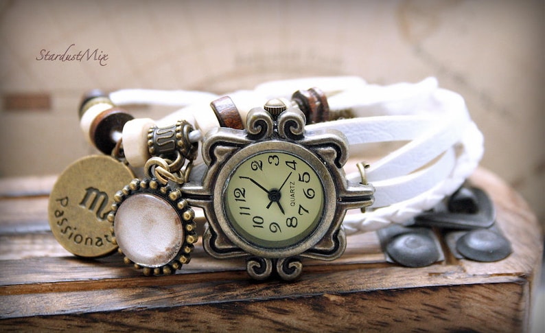 Puede incluir: Un reloj de pulsera de cuero blanco con una esfera redonda de color dorado antiguo. La esfera del reloj tiene marcadores de hora con n&uacute;meros romanos negros y agujas negras. El reloj est&aacute; adornado con un dije que dice "Passionate" y un peque&ntilde;o dije redondo blanco.