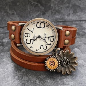 Peut inclure: Une montre-bracelet en cuir marron avec un cadran crème et des chiffres noirs. La montre est ornée d'un charm en forme de tournesol et d'un autre plus petit avec un motif de tournesol. Le bracelet est fixé par des clous métalliques.