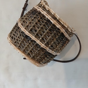May include: A square woven basket with a dark brown leather strap. The basket features a woven pattern in shades of brown and beige. The strap is attached to the top and is adjustable. The basket is designed to be carried over the shoulder, a traditional design.