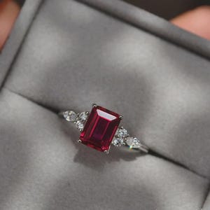 Op de afbeelding: Een zilveren ring met een grote, rechthoekige, dieprode edelsteen. Kleinere, heldere edelstenen flankeren de centrale steen. De ring wordt getoond tegen een grijze achtergrond.