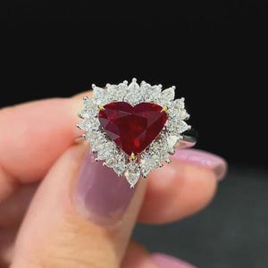 Puede incluir: Un anillo con un rubí en forma de corazón, rodeado por un halo de diamantes blancos. El rubí es de un color rojo intenso, y los diamantes brillan. El anillo está engastado en una banda de plata.