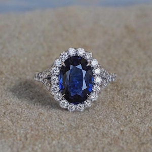 Puede incluir: Un anillo de plata con una gran gema ovalada de color azul oscuro, rodeada por un halo de gemas más pequeñas y claras. El anillo se encuentra sobre una superficie arenosa, mostrando su intrincado diseño y brillo.