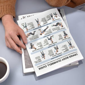 May include: A printed upper body dumbbell guide with exercise illustrations. The guide includes exercises for shoulders, back, chest, and arms. The guide is on top of a stack of magazines, with a cup of coffee in the corner.