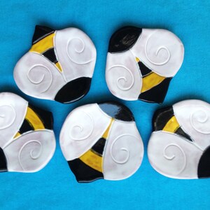 May include: Five ceramic bee-shaped coasters with a white background, black and yellow stripes, and a swirl design.
