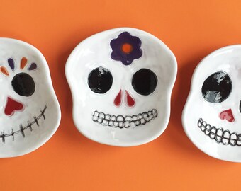May include: Three white ceramic skull-shaped dishes with painted details. The first skull has orange, purple, and black painted details around the eyes and a red nose. The second skull has a purple flower painted on the forehead and black eyes. The third skull has a red heart painted on the forehead and black eyes.