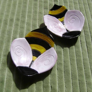 May include: Two ceramic bee-shaped dishes with black and yellow stripes, white wings, and a black head. The dishes are on a green textured surface.