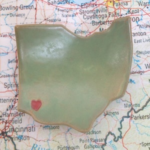 May include: A green ceramic Ohio state outline with a red heart in the center. The outline is sitting on a map of the United States.