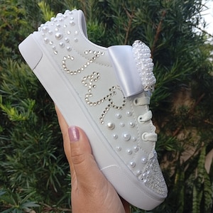 May include: White sneakers adorned with pearls and rhinestones. The shoe features a large bow and the initials "J & R" spelled out in rhinestones. The toe and upper heel are also embellished with rhinestones. The laces are white.