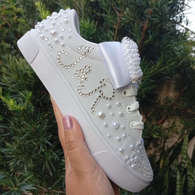 May include: White sneakers adorned with pearls and rhinestones. The shoe features a large bow and the initials "J & R" spelled out in rhinestones. The toe and upper heel are also embellished with rhinestones. The laces are white.