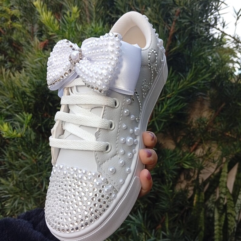 May include: White sneaker with a large, embellished bow and pearl accents. The toe of the shoe is covered in sparkling rhinestones. The shoe has white laces and a white sole. A decorative shoe for special occasions.