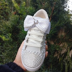 May include: White sneakers with a large, pearl-covered bow and rhinestone accents. The toe of the shoe is covered in small rhinestones. The shoe is held up against a background of green foliage.