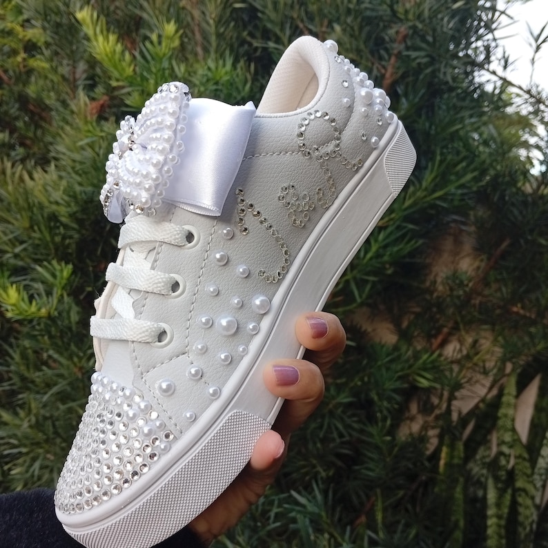 May include: White sneakers adorned with pearls and rhinestones. The shoe features a large bow embellished with pearls on the laces. The toe of the shoe is covered in rhinestones. The shoe is held in front of a green leafy background.
