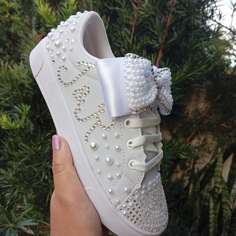 May include: White sneakers adorned with pearls and rhinestones. A large, decorative bow is on the laces. The word "Joy" is spelled out in rhinestones on the side of the shoe. The sneakers have a rubber sole.