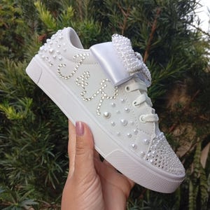 May include: White sneakers adorned with pearls and rhinestones. The shoe features a large bow with pearl embellishments and the word "Love" spelled out in rhinestones. The toe and heel are also covered in rhinestones.
