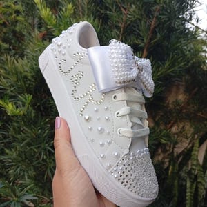 May include: White sneaker embellished with pearls and rhinestones. A large white bow is on the laces. The word "Cute" is spelled out in rhinestones on the side of the shoe. The shoe is held in a hand against a background of green foliage.
