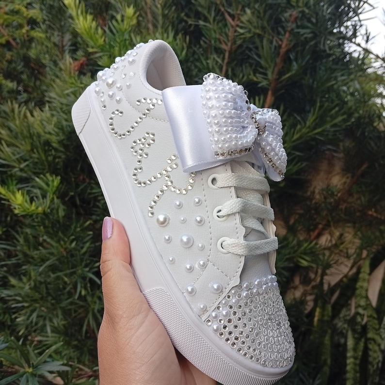 May include: White sneaker embellished with pearls and rhinestones. A large white bow is on the laces. The word "Cute" is spelled out in rhinestones on the side of the shoe. The shoe is held in a hand against a background of green foliage.