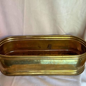 May include: A rectangular brass planter with rounded ends and two curved handles. The planter has a shiny, golden finish and is designed to hold plants or flowers. The interior appears to be empty.