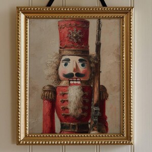 May include: A framed print of a nutcracker soldier in a red uniform and tall hat, holding a rifle. The artwork has a gold frame and is suspended by a black ribbon. The background is a neutral color.