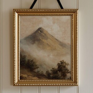 May include: A framed landscape painting depicting a mountain peak, in muted earth tones. The gold frame has a beaded edge, and the artwork is suspended by a black ribbon tied in a bow. The painting evokes a sense of tranquility.