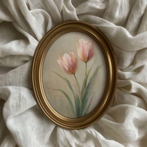 K&ouml;nnte beinhalten: Oval gerahmtes Gem&auml;lde mit zwei rosa Tulpen mit gr&uuml;nen Stielen und Bl&auml;ttern. Das Kunstwerk ist vor einem cremefarbenen Hintergrund platziert und in einem goldfarbenen ovalen Rahmen gerahmt. Das Gem&auml;lde wird auf einem wei&szlig;en, strukturierten Stoff pr&auml;sentiert.