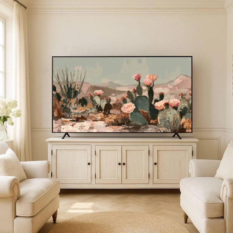 May include: A large flat-screen television displaying a desert landscape painting with cacti and pink flowers. The TV is set on a white cabinet with three sets of doors, in a living room setting with two cream-colored armchairs.