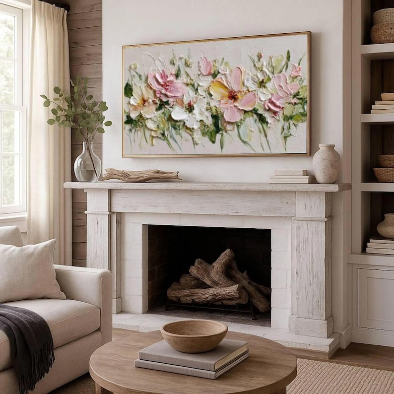 May include: A framed floral painting with a gold border hangs above a white fireplace. The artwork features textured pink, yellow, and white flowers with green foliage. A vase with greenery and a decorative bowl are on the mantel.