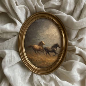 Op de afbeelding: Ovaal ingelijst schilderij met twee paarden die over een veld rennen. De paarden zijn bruin en zwart, tegen een gedempte, bewolkte lucht. De goudkleurige lijst voegt een klassieke touch toe aan het kunstwerk.