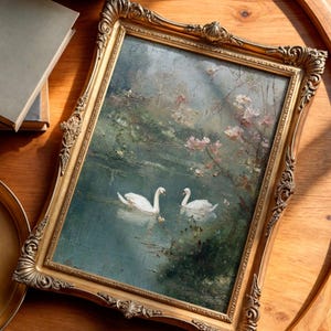 May include: A framed painting featuring two white swans on a lake, surrounded by greenery and pink blossoms. The painting is set in an ornate gold frame, resting on a wooden surface.