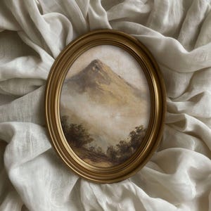 May include: An oval gold-framed painting depicting a mountain scene. The artwork shows a mountain peak in brown and beige tones, with a misty atmosphere. The painting is set against a white, textured background.