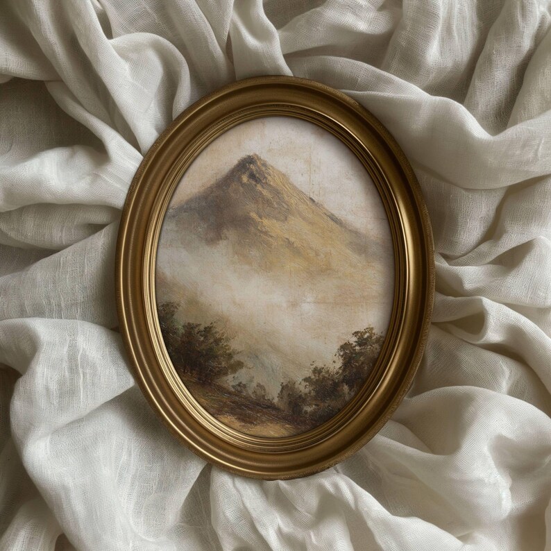 May include: An oval gold-framed painting depicting a mountain scene. The artwork shows a mountain peak in brown and beige tones, with a misty atmosphere. The painting is set against a white, textured background.