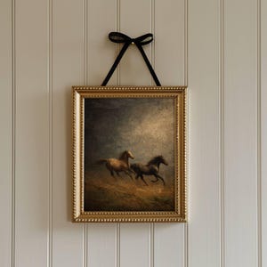 Op de afbeelding: Een ingelijst kunstwerk met twee rennende paarden in een veld, met een goudkleurige, sierlijke lijst. Het kunstwerk wordt opgehangen door een zwart lint dat in een strik is geknoopt. De achtergrond is een neutraal getinte muur.
