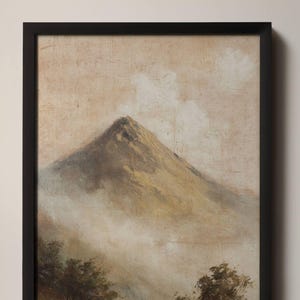 May include: A framed landscape painting depicting a mountain peak in shades of brown and gold, partially obscured by clouds and mist. The artwork is set against a neutral background and framed in a black border. Trees are visible at the bottom.