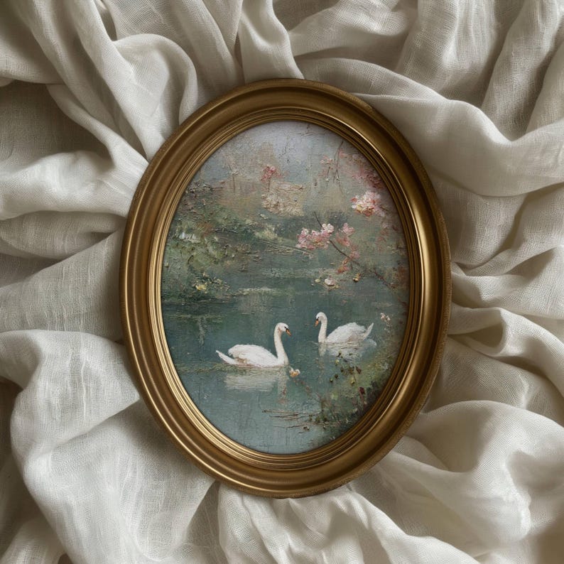 May include: Oval framed painting featuring two white swans on a lake. The artwork has a gold frame and depicts a serene landscape with pink blossoms and green foliage. The painting is set against a soft, white fabric background.