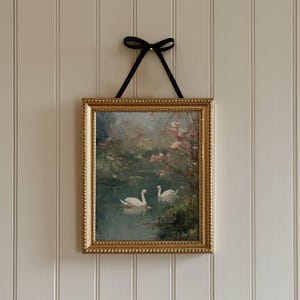 May include: A framed painting of two swans on a lake, set in a gold ornate frame. The artwork is hung by a black ribbon tied in a bow. The painting has a soft, romantic feel with pink blossoms and a muted color palette.