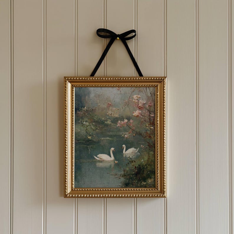 May include: A framed painting of two swans on a lake, set in a gold ornate frame. The artwork is hung by a black ribbon tied in a bow. The painting has a soft, romantic feel with pink blossoms and a muted color palette.