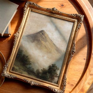 May include: An antique-style framed landscape painting. The artwork depicts a mountain scene, set within an ornate gold-coloured frame. The painting shows a mountain peak in mist, with trees in the foreground. The frame has detailed scrollwork.