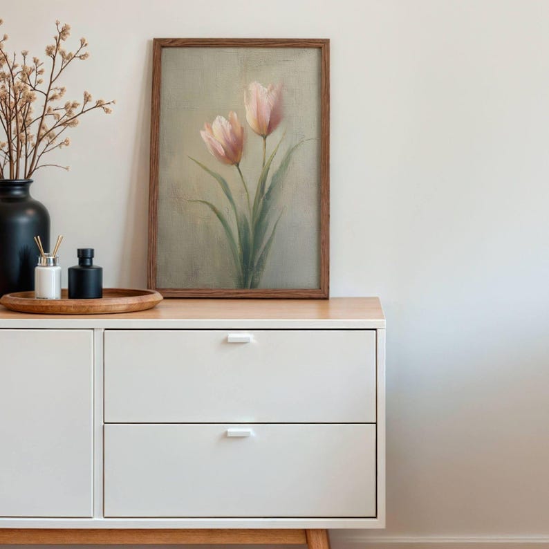 K&ouml;nnte beinhalten: Ein gerahmtes Kunstwerk mit zwei rosa Tulpen mit gr&uuml;nen Stielen und Bl&auml;ttern. Das Gem&auml;lde befindet sich in einem Holzrahmen und steht auf einem wei&szlig;en Schrank mit zwei Schubladen. Eine schwarze Vase und andere dekorative Gegenst&auml;nde sind ebenfalls vorhanden.