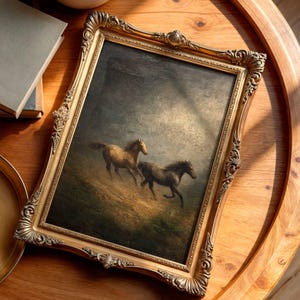 Op de afbeelding: Een ingelijst schilderij met twee paarden die over een grasheuvel rennen onder een bewolkte hemel. Het kunstwerk zit in een gouden, sierlijke lijst. Het schilderij heeft een vintage uitstraling en staat op een houten tafel.