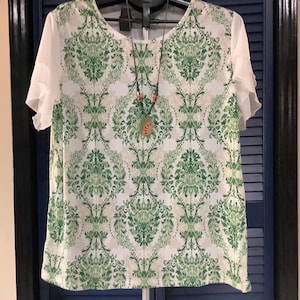 May include: A short-sleeved top featuring a white base with a repeating green damask pattern. The garment has solid white ruffled sleeves and a round neckline. A long necklace with a green pendant and small red beads hangs over the front.