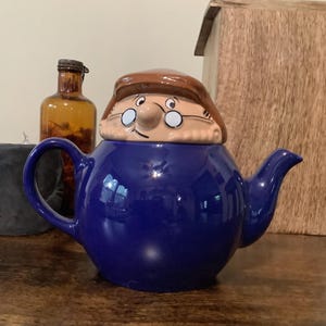 May include: A cobalt blue ceramic teapot with a whimsical face on the lid. The lid features a brown hat and a face with glasses. A brown glass bottle is visible in the background.