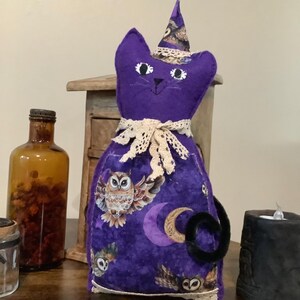 May include: A stuffed fabric cat figure in deep purple, wearing a matching pointed hat and a cream-colored lace collar. The body features a printed pattern of owls and crescent moons. The cat has stitched facial features and a black pipe cleaner tail.