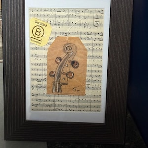 May include: Framed artwork featuring sheet music as a backdrop. A drawing of a violin scroll is on a brown paper tag. A yellow tag with the text "Certified B Corporation" is attached. The frame is dark brown.