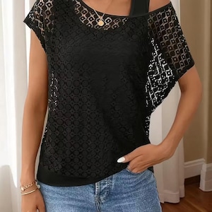 Puede incluir: Top de encaje negro con diseño asimétrico. El top presenta un cuello redondo y una manga corta. El intrincado patrón de encaje añade un toque delicado a la prenda. El top se combina con unos vaqueros azules.