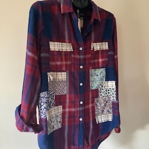 May include: A long-sleeve button-down shirt in a red and blue plaid pattern. The shirt features a collar, a front button placket, and multiple rectangular fabric patches with various floral and checkered prints sewn onto the front panels.