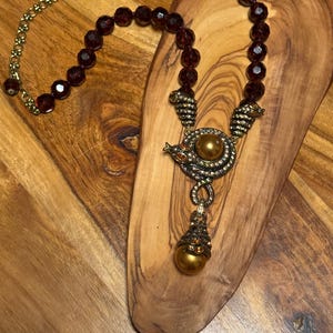 May include: A necklace featuring dark red faceted beads and a gold-toned metal pendant. The pendant is shaped like a coiled creature with small amber-colored crystals and a large, smooth, metallic gold sphere at the center and bottom.