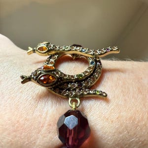 Könnte beinhalten: Ein goldfarbener Armbandverschluss in Form von zwei Schlangen, verziert mit bunten Strasssteinen. Der Verschluss hat einen großen, facettierten, dunkelvioletten Perlenanhänger. Die Augen der Schlangen sind orange, und das Design ist aufwendig und detailliert.
